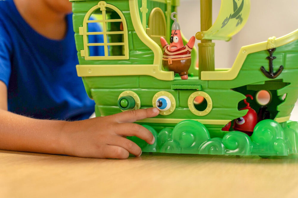 Zestaw figurek FUNRISE SpongeBob Latający Holender 73110 Zbliżenie na dłoń dziecka wsuwającą wystające oko w burtę zabawkowego statku pirackiego SpongeBoba. Figurka Patryka Rozgwiazdy w pirackim stroju stoi na krawędzi powyżej, prezentując interaktywne funkcje zestawu Latający Holender.