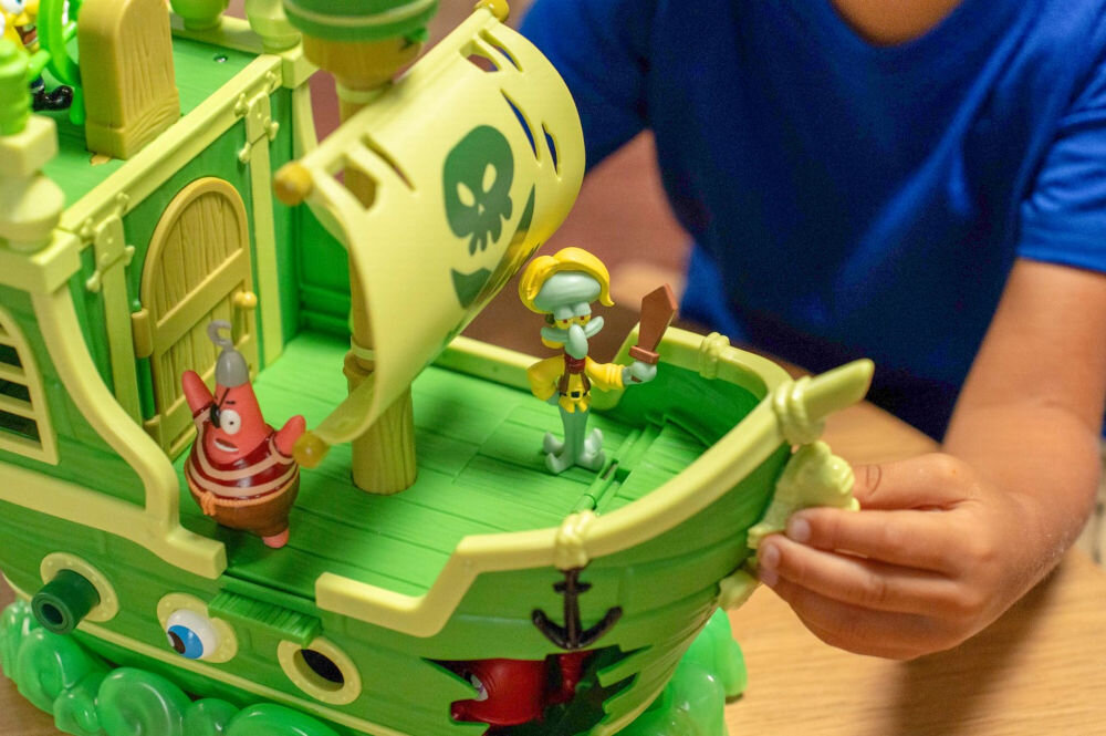 Zestaw figurek FUNRISE SpongeBob Latający Holender 73110 Zbliżenie na przedni pokład zielonego statku pirackiego z figurką Skalmara Obłego ubranego w piracki strój z mieczem. W tle widoczny jest żółty żagiel z pirackim symbolem oraz figurka Patryka.