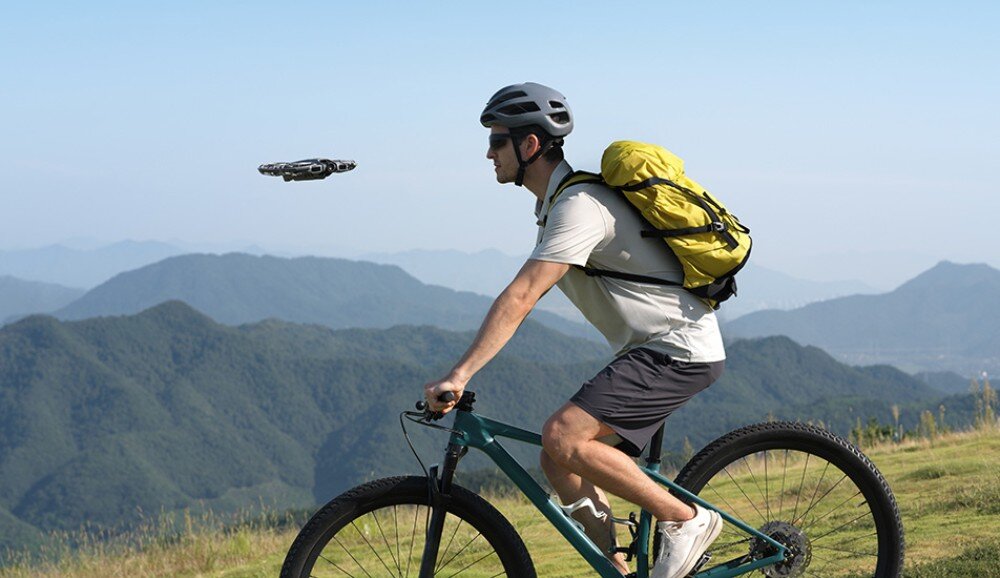 Dron DJI Neo 2 Fly More Combo Rowerzysta w kasku i z żółtym plecakiem jedzie po trawie w górach, a obok niego leci mały dron, śledzenie obiektów, funkcje