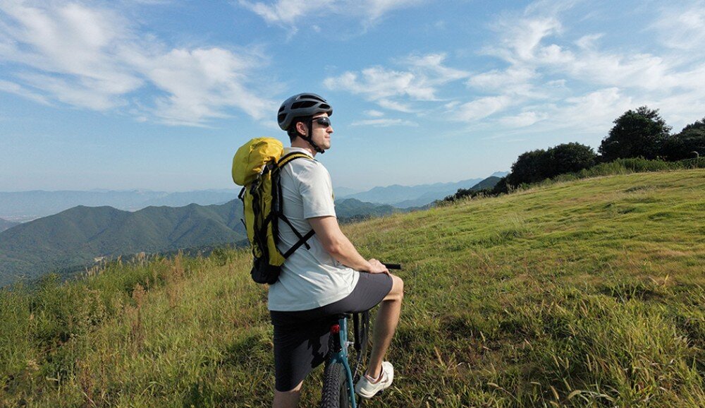 Dron DJI Neo 2 Fly More Combo Rowerzysta w kasku stoi na wzgórzu z widokiem na górskie pasma pod niebem z chmurami, tryby nagrywania