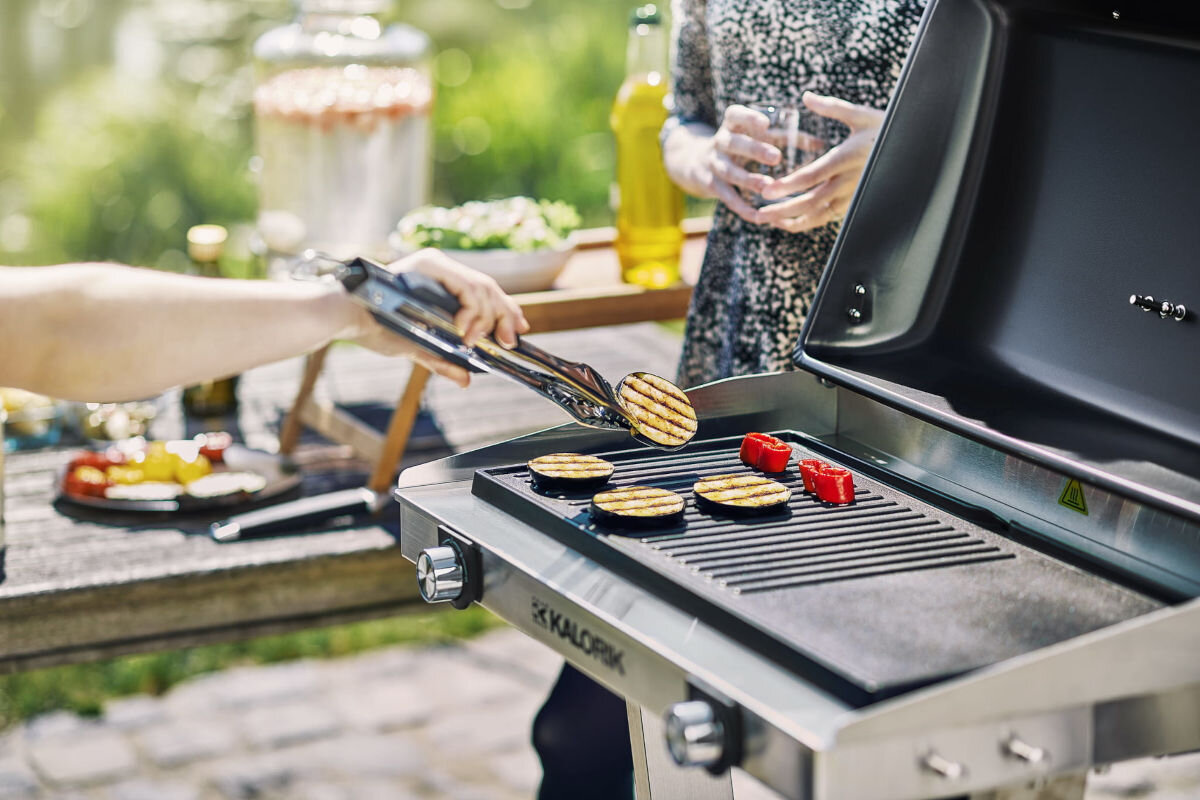 Grill elektryczny KALORIK TKG EBBQ 1002 L, płyta, grzewcza, jednoczesne, potrawy, osoba operuje szczypcami i potrawą na ruszcie grilla