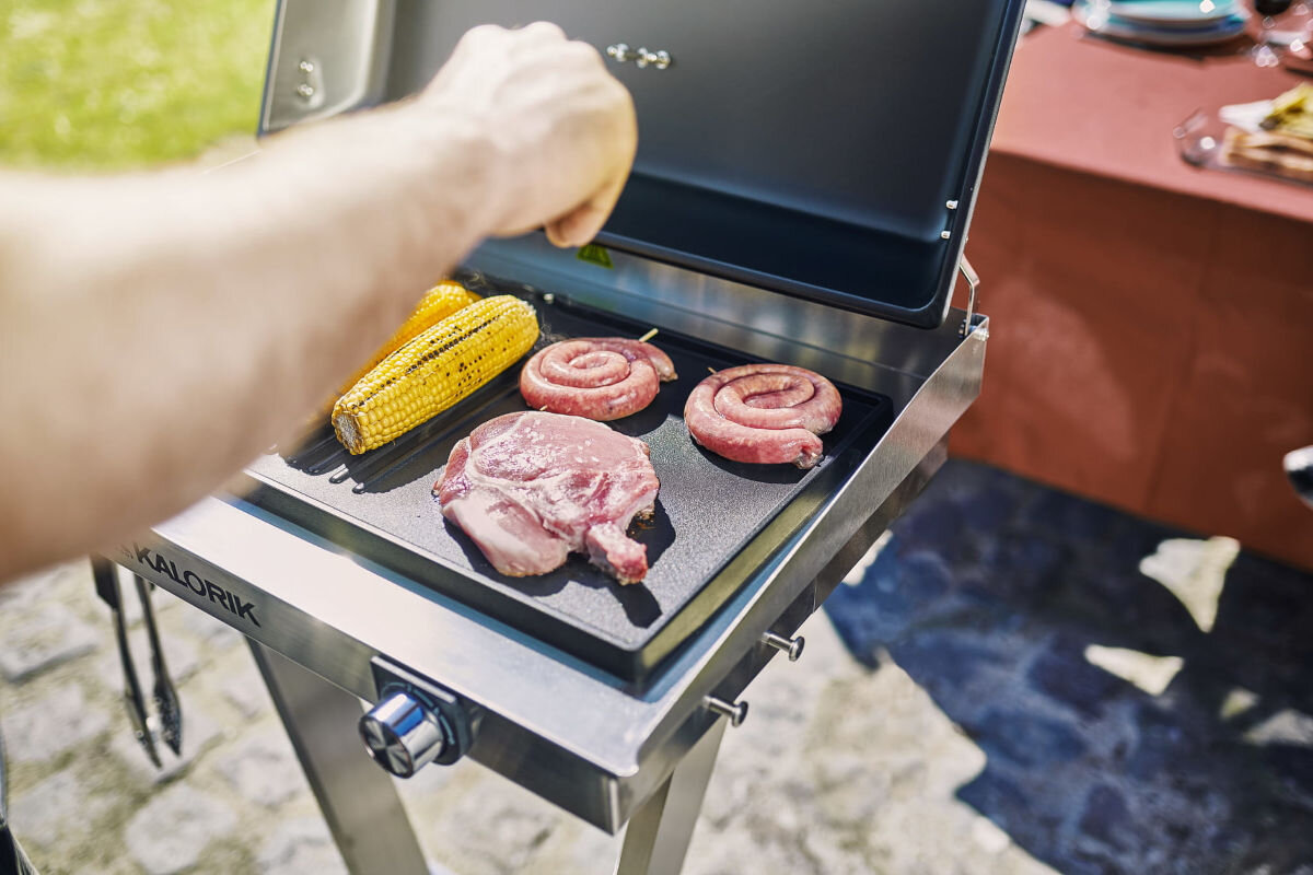 Grill elektryczny KALORIK TKG EBBQ 1002 L, taca, ociekanie, pokrywa, rozpryskiwanie, osoba operuje nad rusztem grilla