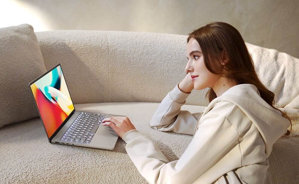 Kobieta w beżowej bluzie używająca laptopa ASUS ZenBook z kolorowym ekranem OLED siedząca na jasnej sofie w przytulnym wnętrzu domowym - 14-calowy ekran FHD+ 