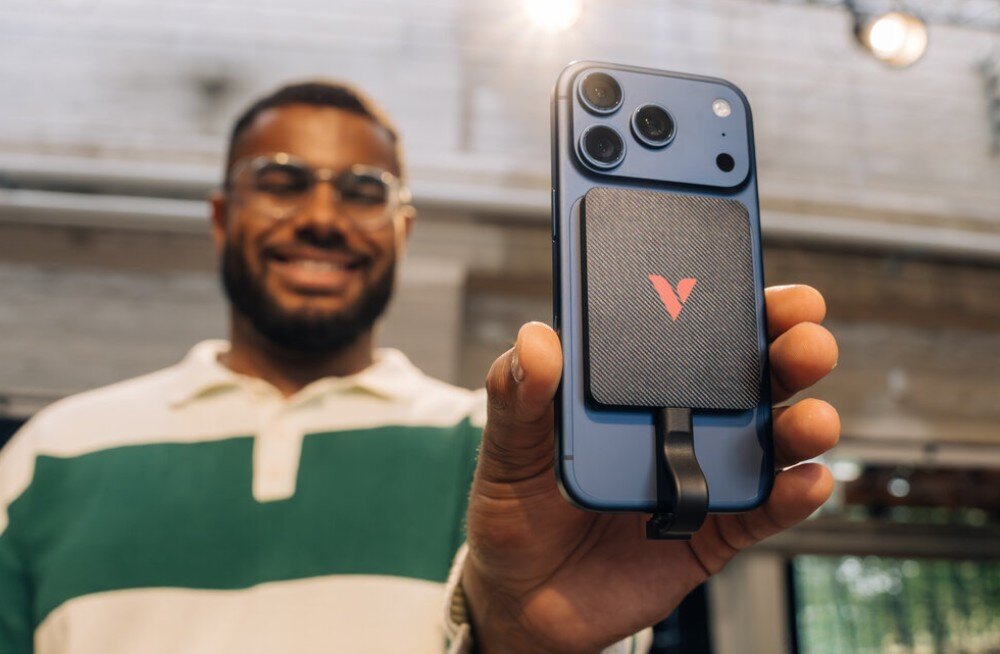 Dysk VERBATIM SnapBack Ultra Slim Uśmiechnięty mężczyzna w okularach prezentuje tył niebieskiego smartfona, do którego przymocowany jest czarny kwadratowy dysk z czerwonym logo 'V' oraz krótkim kablem podłączonym do portu, opis