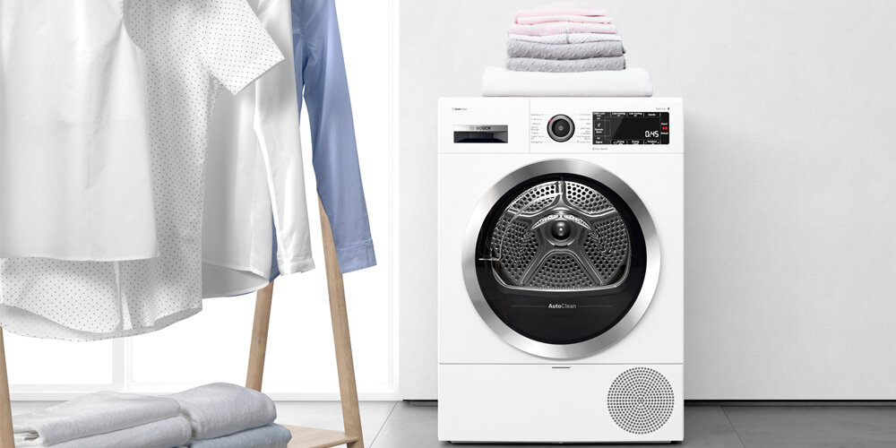 SUSZARKA BOSCH WQG244CSPL Na zdjęciu widoczna jest suszarka Bosch z bębnem z charakterystyczną metalową perforacją. Na urządzeniu leżą starannie złożone ręczniki w jasnych kolorach. Po lewej stronie znajduje się stojak z powieszonymi koszulami i bluzkami. Przestrzeń jest jasna, czysta i minimalistyczna – prezentuje pralnię lub garderobę IronAssist mniej zagnieceń