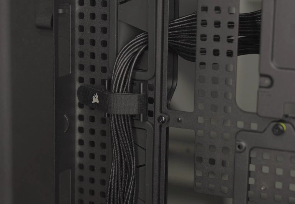 Obudowa CORSAIR Frame 5000D RS ARGB Widok na tył tacki płyty głównej z systemem zarządzania okablowaniem. Wiązka czarnych przewodów w oplocie jest uporządkowana i przytrzymywana przez materiałowy rzep z logo producenta, przechodząc przez otwory w konstrukcji