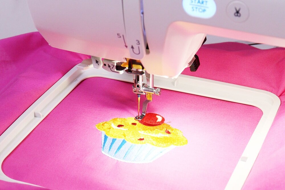 Hafciarka wykonuje wzór babeczki na różowym materiale. Na maszynie widoczny jest przycisk z napisem \'START STOP\'.