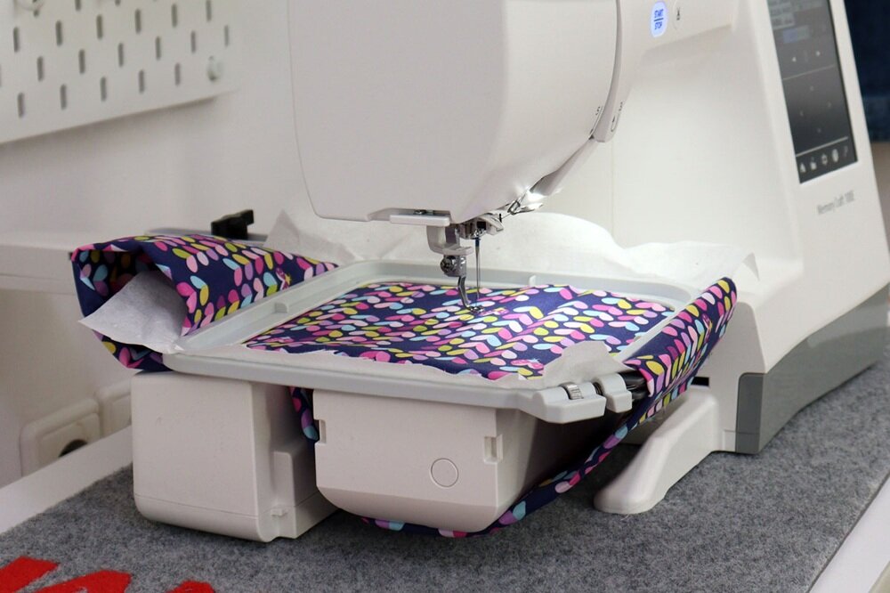 Hafciarka z białą obudową pracuje nad kolorowym materiałem w ramce. Na obudowie widoczny jest napis \'Memory Craft 100E\'.