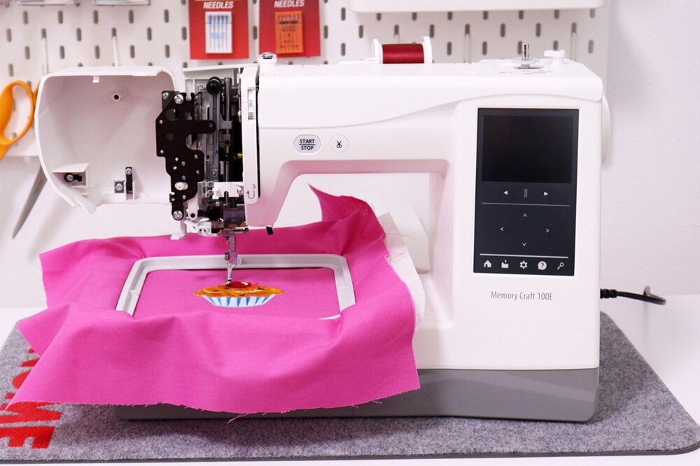 Hafciarka z wyświetlaczem i panelem sterowania, na której znajduje się różowy materiał z haftowanym wzorem. Na maszynie widoczny jest napis \'Memory Craft 100E\'.