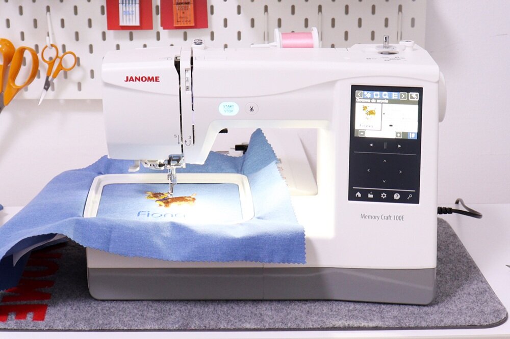 Hafciarka z logo JANOME, model Memory Craft 100E, z wyświetlaczem i materiałem w ramce do haftowania. W tle widoczne są nożyczki i inne akcesoria krawieckie na ścianie.