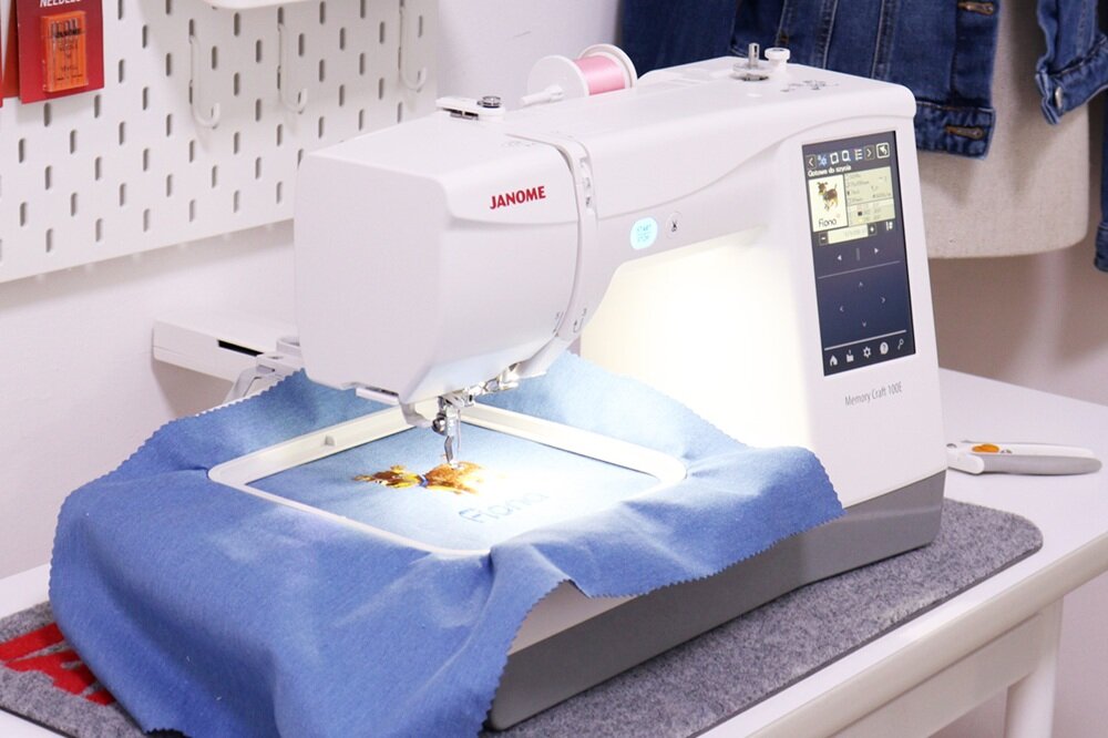 Na zdjęciu widoczna jest hafciarka z logo \'JANOME\', pracująca nad niebieskim materiałem. Na ekranie urządzenia wyświetlane są opcje sterowania i wzór haftu.