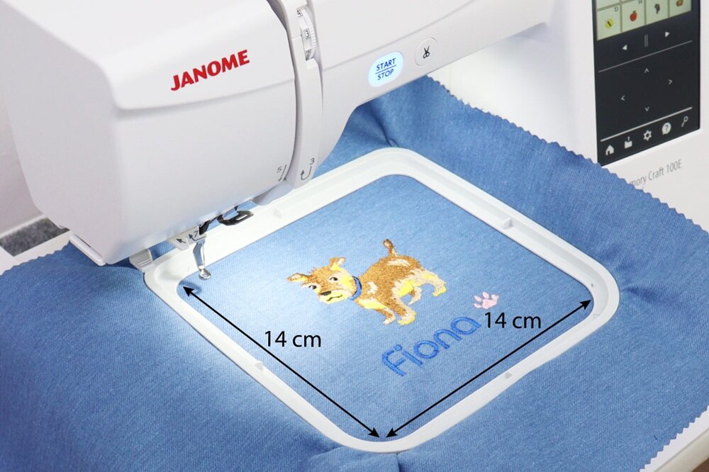 Hafciarka z logo JANOME wykonuje haft przedstawiający psa i napis \'Fiona\' na niebieskim materiale. Obszar haftu ma wymiary 14 cm na 14 cm.