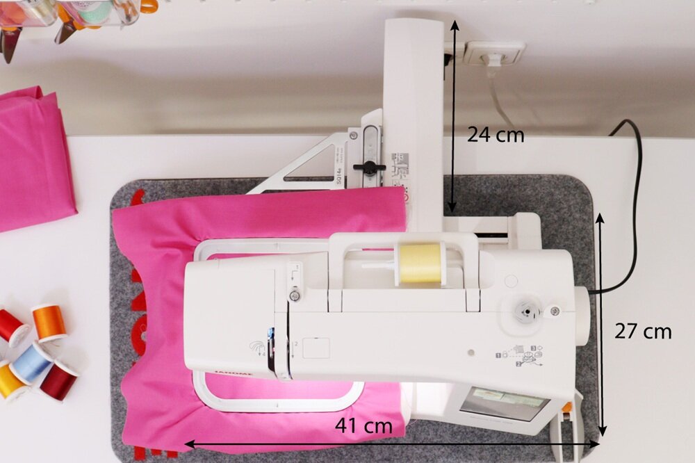 Na obrazie widoczna jest hafciarka z różowym materiałem, z wymiarami 41 cm, 24 cm i 27 cm. Obok znajdują się kolorowe nici i różowy materiał.