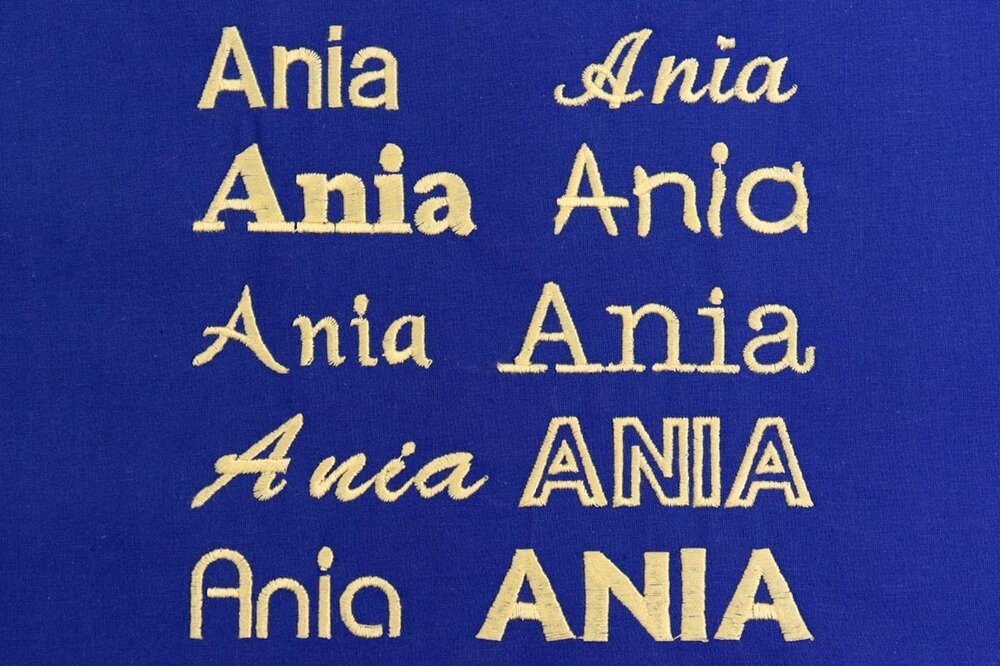 Na niebieskim tle znajduje się wyhaftowane słowo \'Ania\' w różnych stylach czcionek, wszystkie w kolorze złotym.