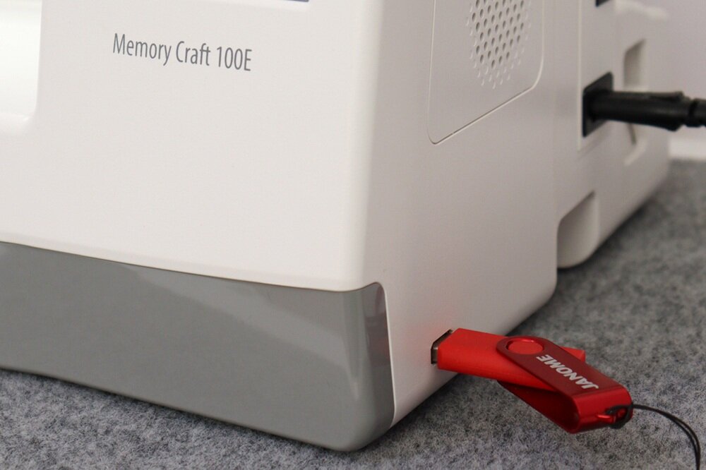 Biała maszyna z napisem \'Memory Craft 100E\' na obudowie. Do urządzenia podłączony jest czerwony pendrive z napisem \'JANOME\'.