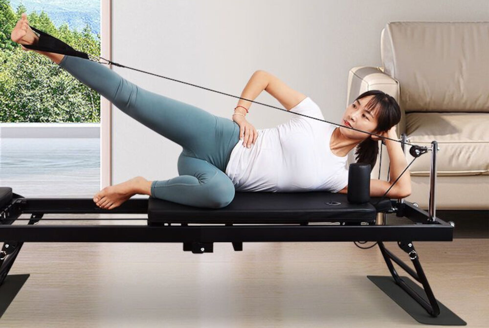 Reformer do pilatesu EXTRALINK Lifestyle PB1001 Czarny pokój osoba ćwiczy na reformerze w tle okno kanapa zagłówek z dwustopniową regulacją dopasowanie ułożenia głowy do rodzaju ćwiczeń i indywidualnych preferencji odciąża odcinek szyjny kręgosłupa sprzyja zachowaniu prawidłowej postawy i zwiększa komfort