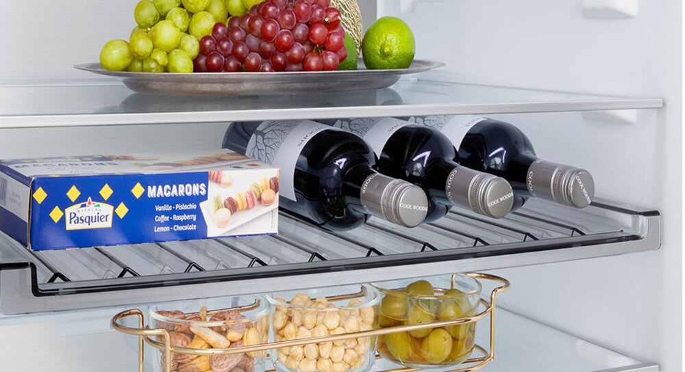 LODÓWKA LG GBBW726AEV Wnętrze lodówki LG z metalową półką na wino, na której leżą trzy butelki w pozycji poziomej obok pudełka z makaronikami. Nad nią ustawiono talerz z winogronami i cytrusami, a poniżej złotą tacę z przekąskami i oliwkami, co podkreśla praktyczną półkę na butelki i elegancką organizację przestrzeni