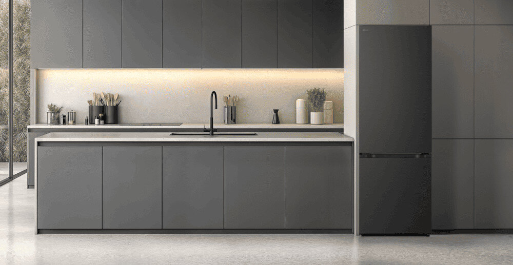 LODÓWKA LG GBBW322CEV Szara zabudowa kuchenna z długim blatem, wyspą i czarną baterią pośrodku, po prawej stronie stoi wolnostojąca lodówko-zamrażarka w grafitowym kolorze, a całość oświetla delikatne podświetlenie LED pod górnymi szafkami