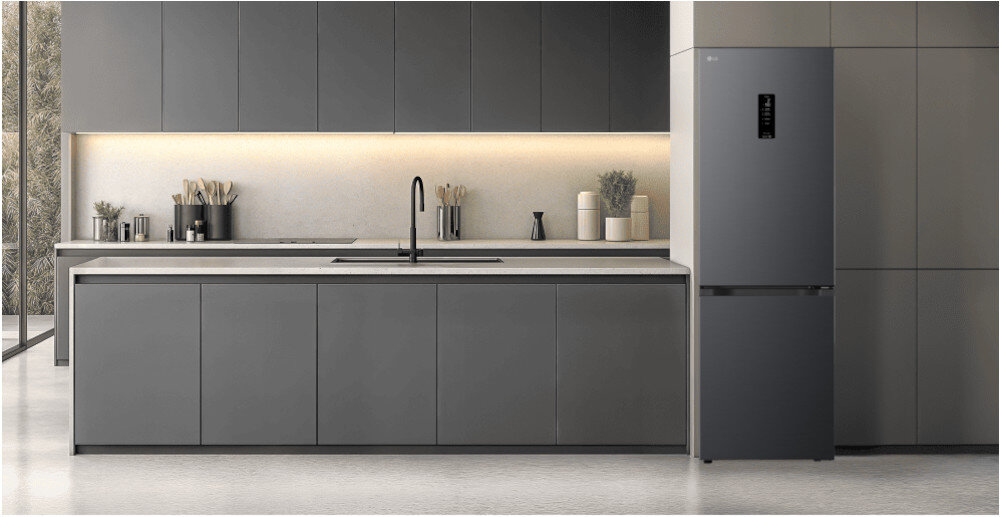 LODÓWKA LG GBBSJ11DEP Minimalistyczna kuchnia w tonacji czarnej z długą wyspą i gładkimi frontami bez uchwytów, oświetlona subtelną listwą LED pod szafkami wiszącymi. Po prawej stronie zabudowy stoi wolnostojąca lodówka w kolorze dopasowanym do mebli, tworząc spójną, elegancką aranżację