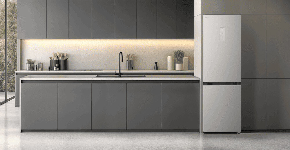 LODÓWKA LG GBBS525CPY Minimalistyczna kuchnia w tonacji grafitowej z długą wyspą i gładkimi frontami bez uchwytów, oświetlona subtelną listwą LED pod szafkami wiszącymi. Po prawej stronie zabudowy stoi wolnostojąca lodówka w kolorze dopasowanym do mebli, tworząc spójną, elegancką aranżację