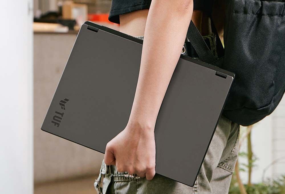 Osoba niesie zamknięty laptop ASUS TUF Gaming z aluminiową pokrywą, podkreślając mobilność w codziennym użytkowaniu – aluminiowy panel dolny o 27% lżejszy i 72% cieńszy od plastiku z naturalnym odprowadzaniem ciepła i pełną sztywnością mechaniczną