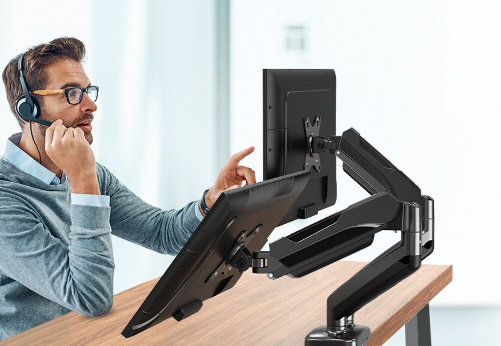 Uchwyt do monitora REDRAGON GMK915 Mężczyzna w słuchawkach z mikrofonem siedzi przy biurku i wskazuje palcem na jeden z dwóch monitorów zamontowanych na czarnym, podwójnym uchwycie przymocowanym do krawędzi drewnianego blatu