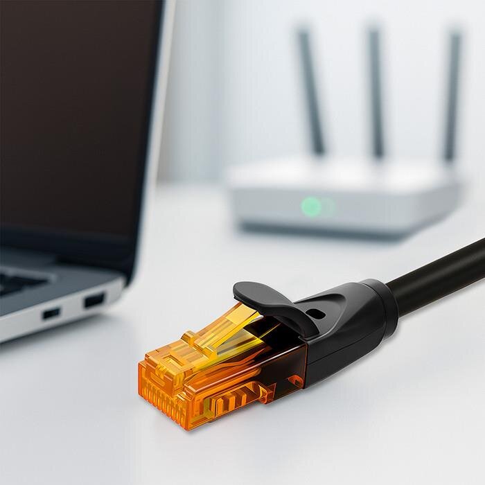 Na obrazie widoczny jest czarny kabel sieciowy z pomarańczową wtyczką RJ-45, leżący obok laptopa i routera z trzema antenami.