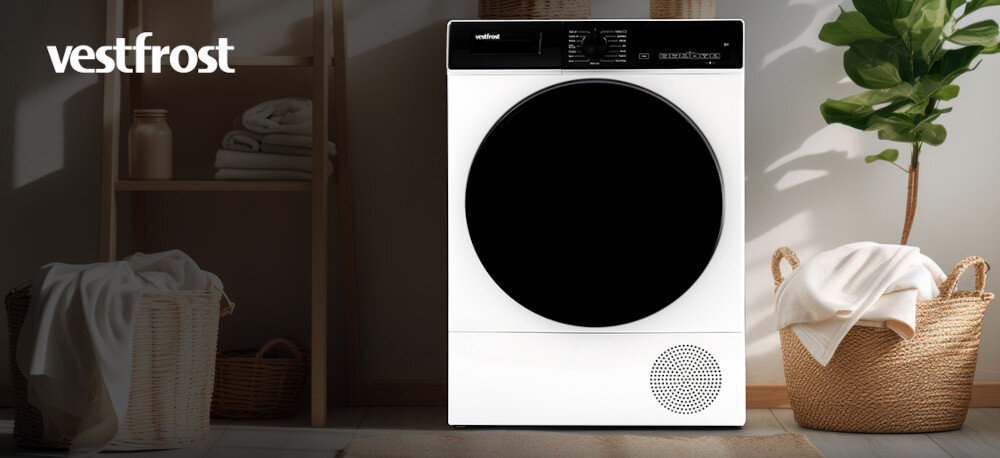 Suszarka VESTFROST VT-FW4EHY70EU Na zdjęciu widoczna jest suszarka bębnowa Vestfrost o ciemnym froncie, ustawiona w stylowym, jasnym pomieszczeniu. Po bokach stoją wiklinowe kosze z ręcznikami, a wnętrze jest uporządkowane i dobrze oświetlone naturalnym światłem