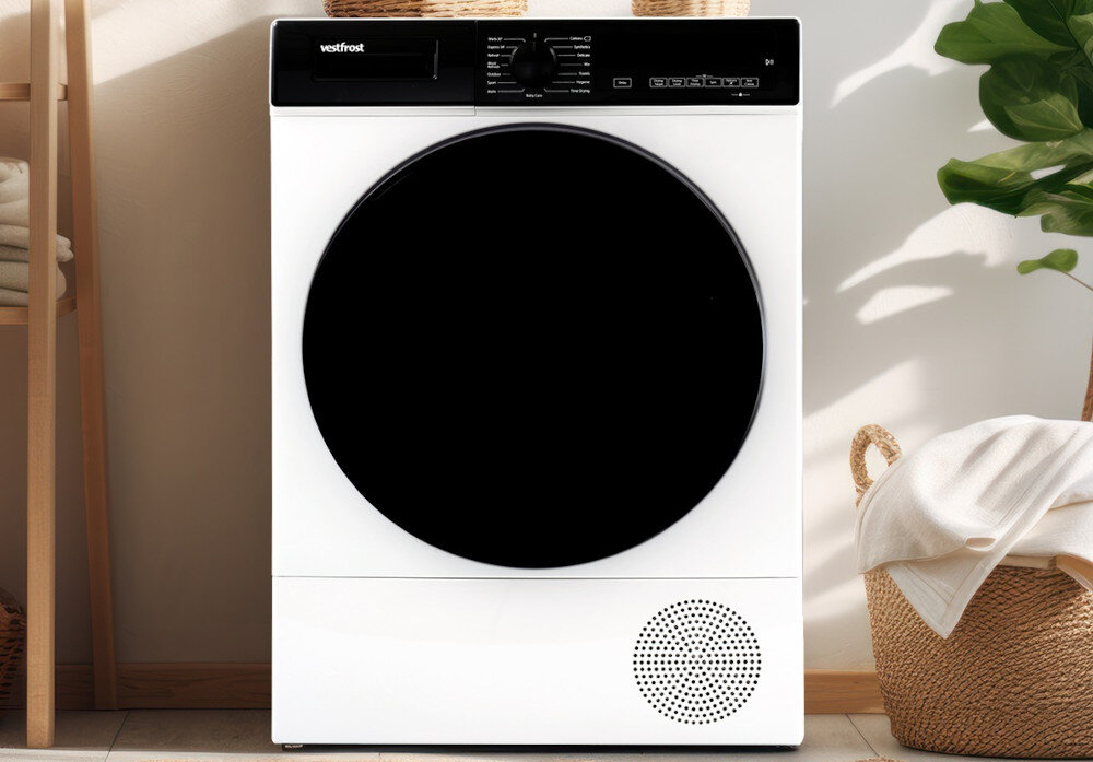 Suszarka VESTFROST VT-FW4EHY70EU Na zdjęciu znajduje się suszarka bębnowa Vestfrost w białym kolorze, umieszczona w jasnym, eleganckim wnętrzu. Urządzenie stoi pomiędzy półką z ręcznikami a koszem, a obok widoczna jest roślina doniczkowa, co podkreśla przytulny charakter aranżacji suszarka do domu elegancka suszarka z wyświetlaczem suszarka premium