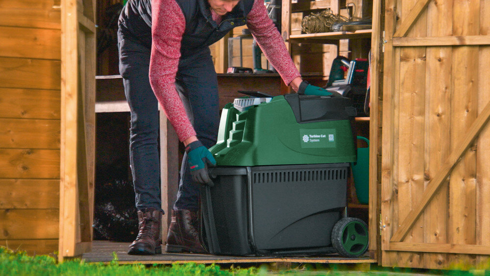 Rozdrabniacz do gałęzi BOSCH AXT 25 TC elektryczny 2500 W sekator + rękawice garaż szopa drewniana konstrukcja rozdrabniacz ustawiony pod ścianą osoba wyciąga rozdrabniacz z szopy dwa praktyczne kółka wygodny uchwyt transportowy można łatwo zmienić miejsce pracy bez wysiłku waga około 30,5 kg komfort i bezpieczeństwo