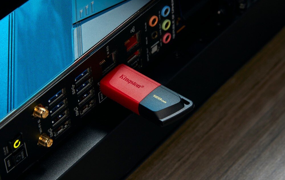 Czarno-czerwony pendrive z napisem \'Kingston\' i \'128GB\' jest podłączony do portu USB w komputerze. W tle widoczne są różne złącza i porty na obudowie komputera.