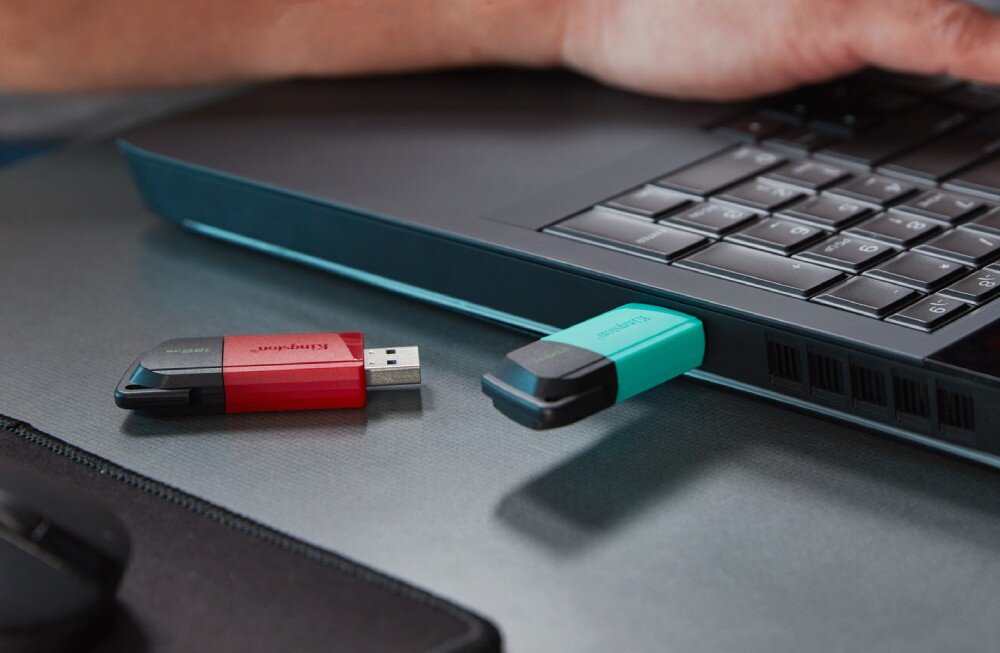 Na obrazie widać dwa pendrive\'y, jeden czerwono-czarny z napisem \'Kingston\', a drugi turkusowo-czarny, podłączone do laptopa. Obok znajduje się ręka na klawiaturze laptopa.
