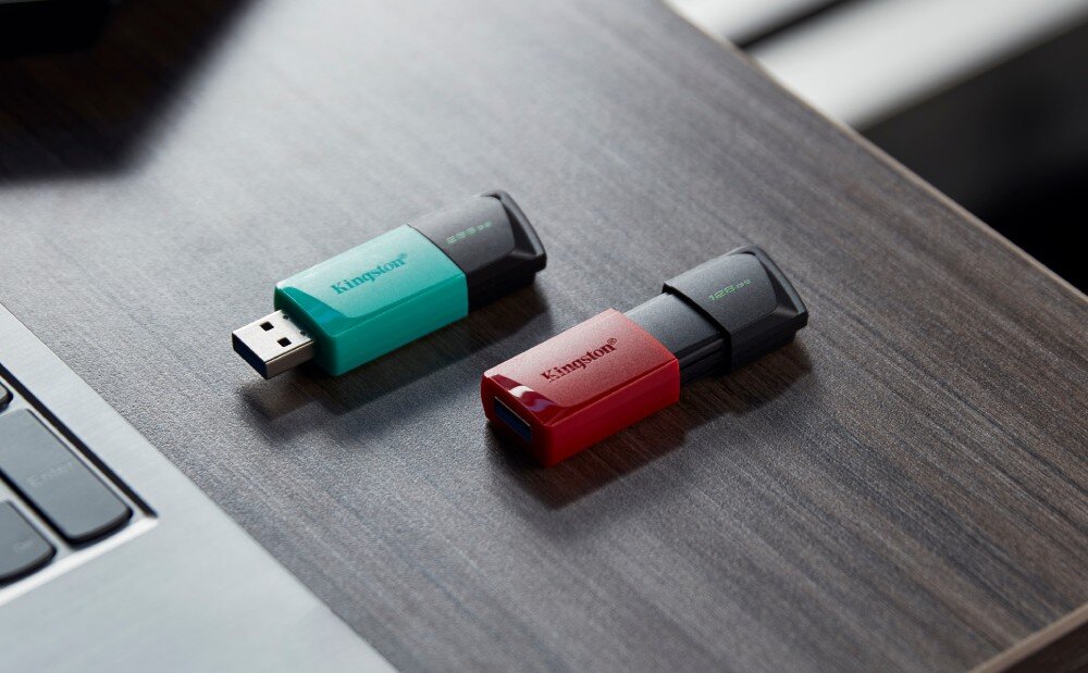 Na drewnianym blacie leżą dwa pendrive\'y, jeden w kolorze turkusowym z napisem \'Kingston\', drugi w kolorze czerwonym również z napisem \'Kingston\'. Oba mają czarne zatyczki i są obok laptopa.