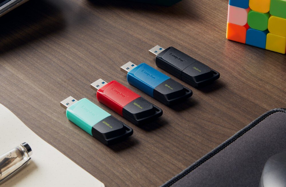 Na drewnianym biurku leżą cztery pendrive\'y w różnych kolorach: miętowym, czerwonym, niebieskim i czarnym. Obok znajduje się kolorowa kostka i fragment notatnika.