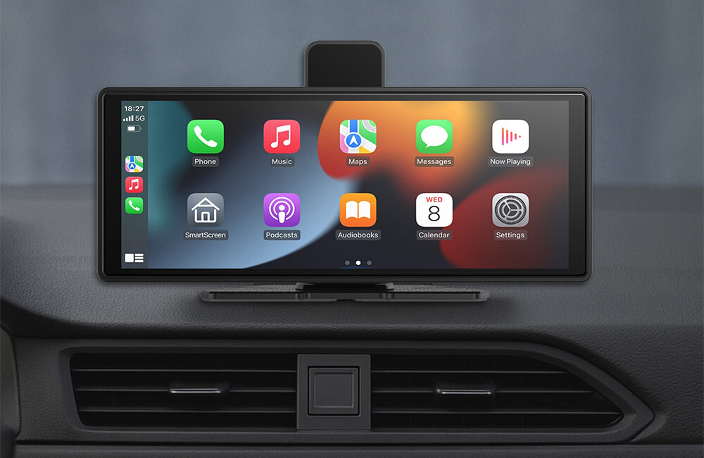 Na obrazie widoczny jest ekran z interfejsem CarPlay zamontowany na desce rozdzielczej samochodu. Na ekranie wyświetlane są ikony aplikacji takich jak Phone, Music, Maps, Messages, Now Playing, SmartScreen, Podcasts, Audiobooks, Calendar i Settings.