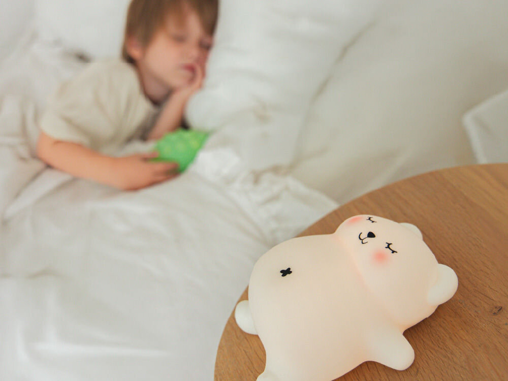 Na drewnianym stoliku znajduje się lampka nocna w kształcie misia z zamkniętymi oczami i uśmiechem. W tle widać dziecko śpiące w łóżku.