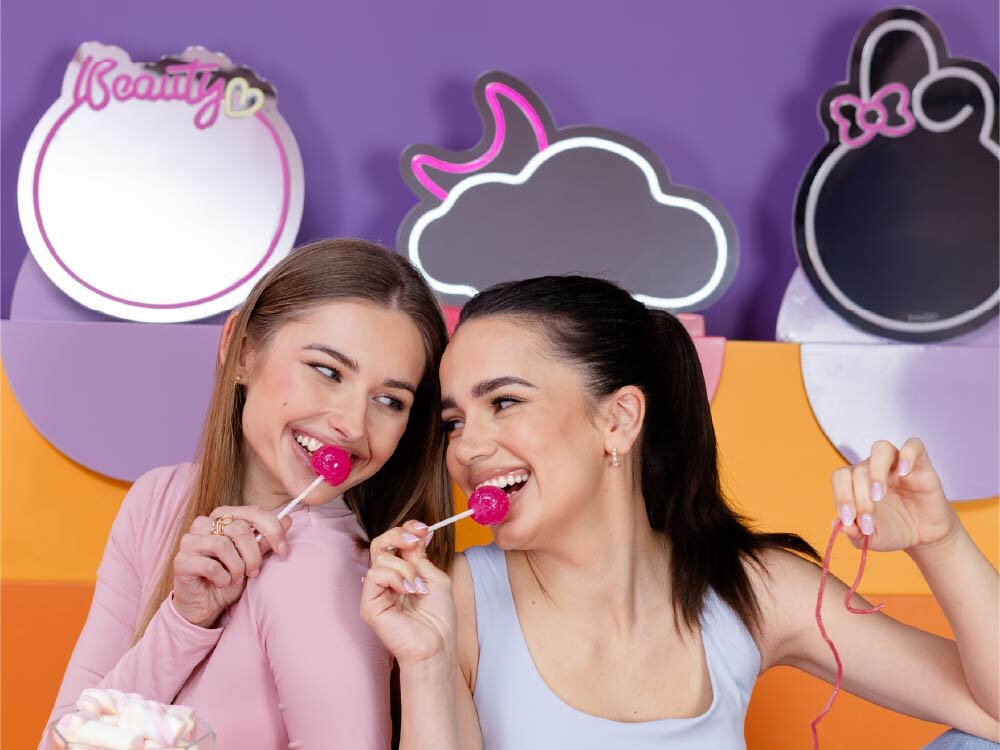 Dwie osoby trzymające lizaki, uśmiechają się do siebie na tle kolorowych luster w kształcie chmurki i innych form. Na jednym z luster widoczny jest napis \'Beauty\'.