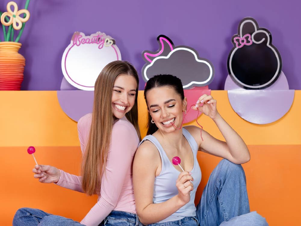 Dwie osoby siedzące na tle kolorowej ściany z lusterkami o różnych kształtach, trzymające lizaki. Na jednym z lusterek widoczny jest napis \'Beauty\'.