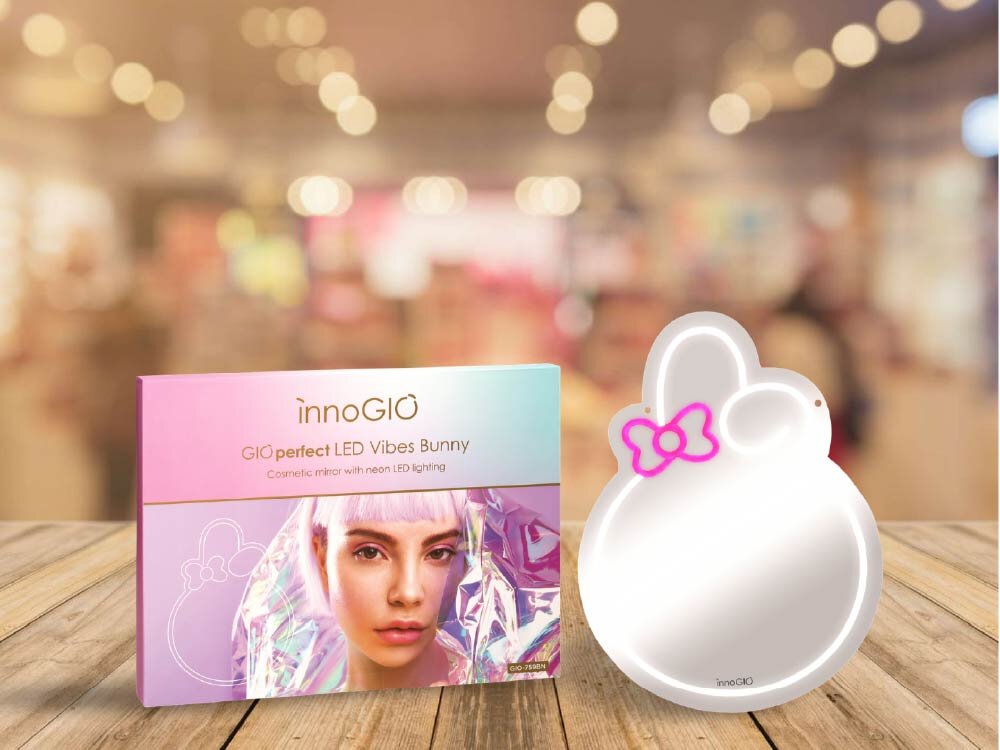 Na drewnianym stole znajduje się opakowanie z napisem \'innoGIO GIOperfect LED Vibes Bunny Cosmetic mirror with neon LED lighting\' oraz lusterko w kształcie królika z różową kokardką. Tło jest rozmyte, przypominające wnętrze sklepu media expert.