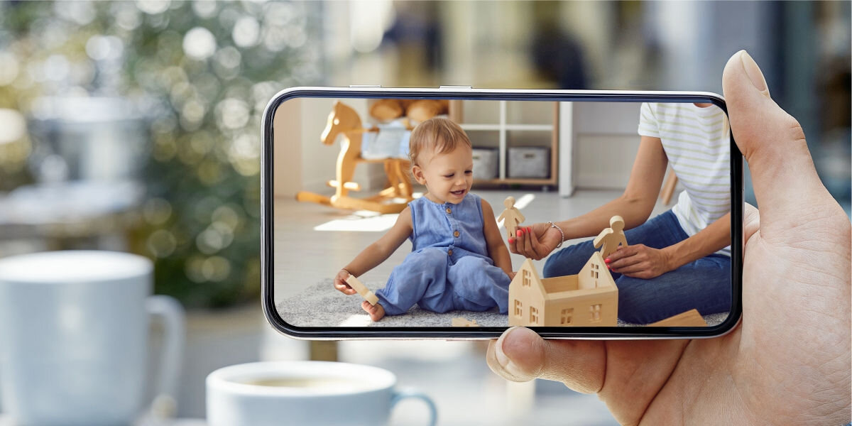 Osoba trzyma smartfon, na którym wyświetlany jest obraz dziecka bawiącego się drewnianymi zabawkami. W tle widoczne są filiżanki i rozmyte elementy wnętrza.