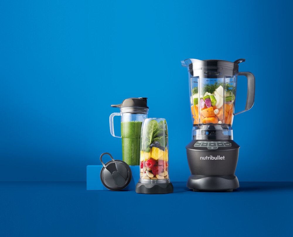 Na niebieskim tle znajduje się blender kielichowy z logo \'nutribullet\', wypełniony różnorodnymi warzywami i owocami. Obok stoją dwa mniejsze pojemniki, jeden z zielonym płynem, drugi z kolorowymi owocami, oraz czarny pokrywka.