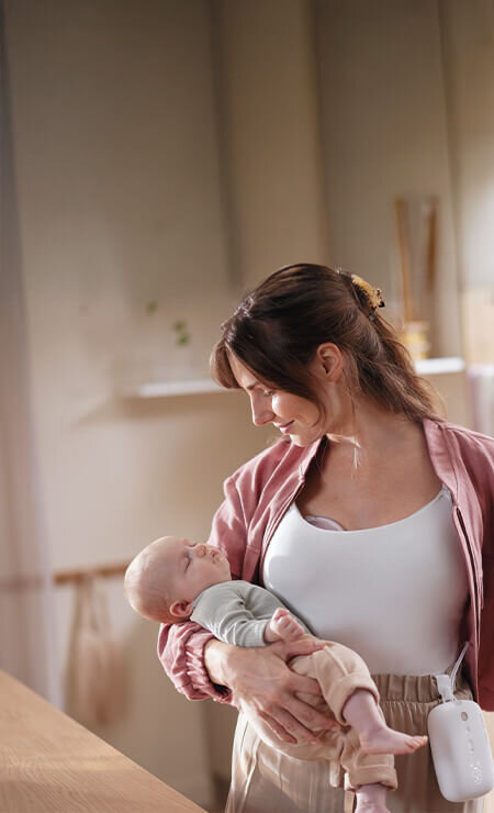 Zdjęcie przedstawiającą kobietę, która używa laktatora. Na prawym ręku trzyma niemowlaka, a w lewej dłoni szklanki.