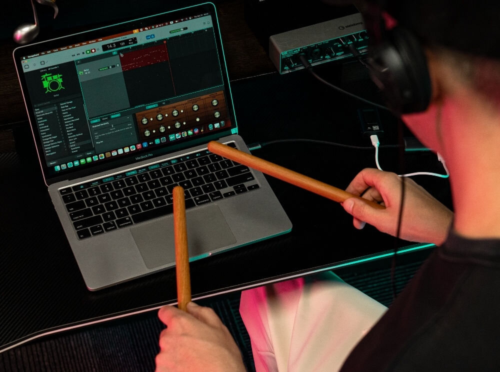  Przenośna perkusja elektroniczna AeroBand PocketDrum 2 Max – Bluetooth, velocity, tryby perkusyjne, osoba z założonymi słuchawkami nausznymi oraz pałeczkami od perkusji siedząca przy laptopie