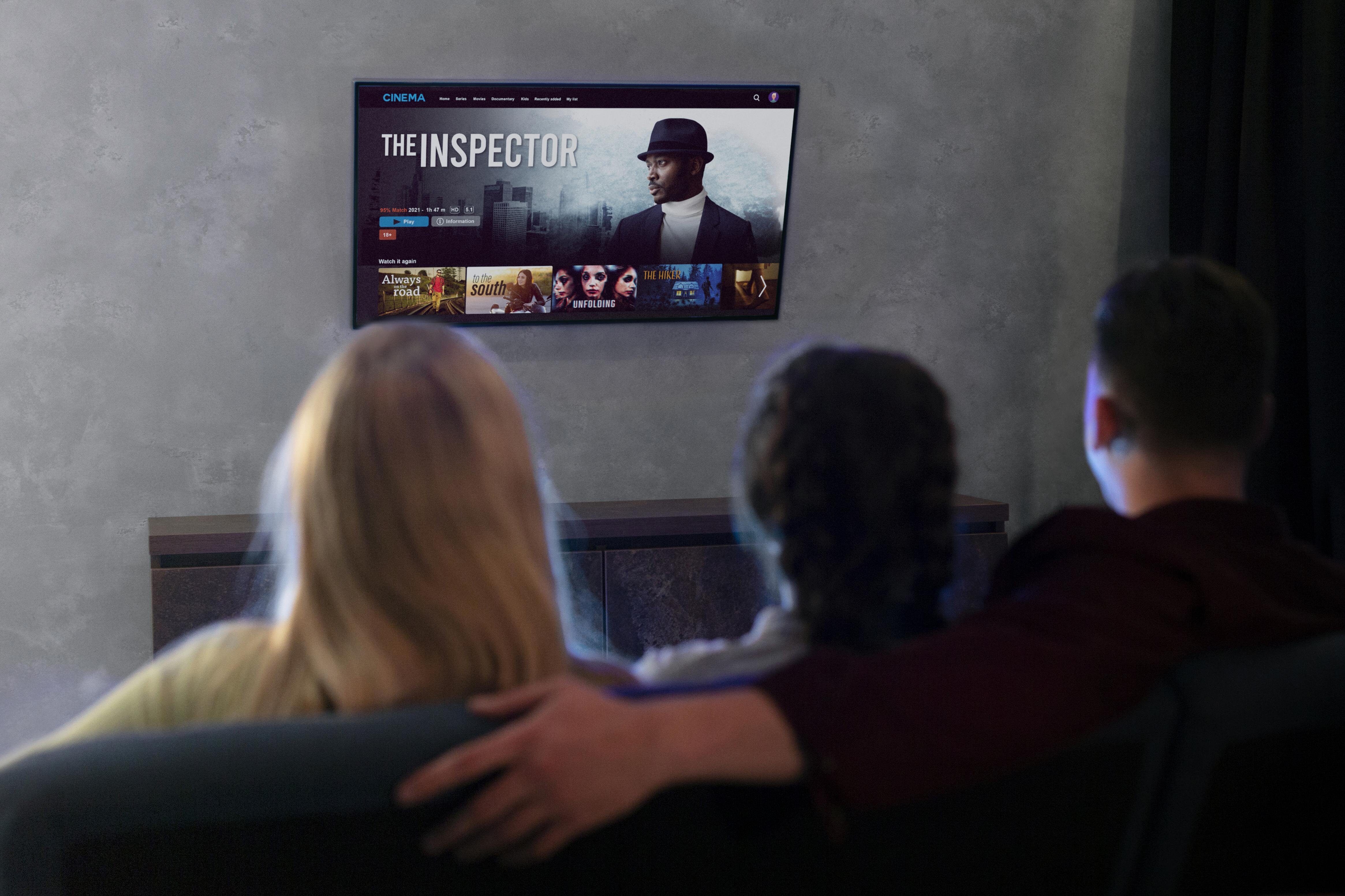 Na ścianie wisi telewizor, na którym wyświetlany jest ekran z napisem \'THE INSPECTOR\'. Przed telewizorem siedzą trzy osoby na kanapie, oglądając program.