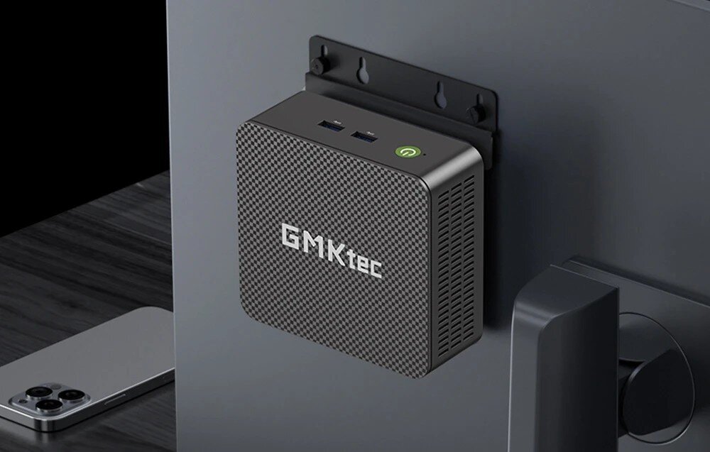 Komputer GMKTEC G3S Małe kwadratowe urządzenie o fakturze przypominającej karbon zamontowane na tylnej obudowie monitora za pomocą czarnego uchwytu