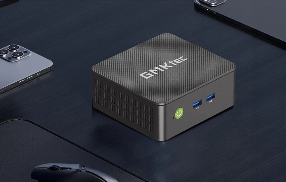 Komputer GMKTEC G3S Kwadratowe urządzenie o ciemnej obudowie z napisem 'GMKtec' leżące na czarnym biurku obok smartfona i myszki komputerowej