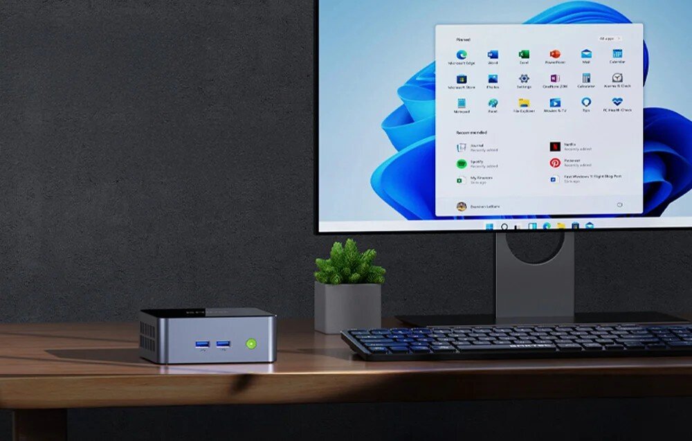 Komputer GMKtec M2 Pro S Mały komputer z zielonym przyciskiem zasilania stojący na drewnianym biurku obok klawiatury i monitora wyświetlającego menu start systemu