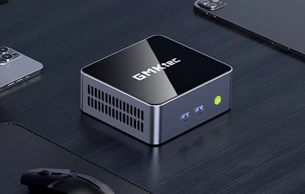 Komputer GMKtec M2 Pro S Ciemne, kwadratowe urządzenie z podświetlonym zielonym przyciskiem zasilania i dwoma portami USB na przednim panelu, leżące na czarnym biurku