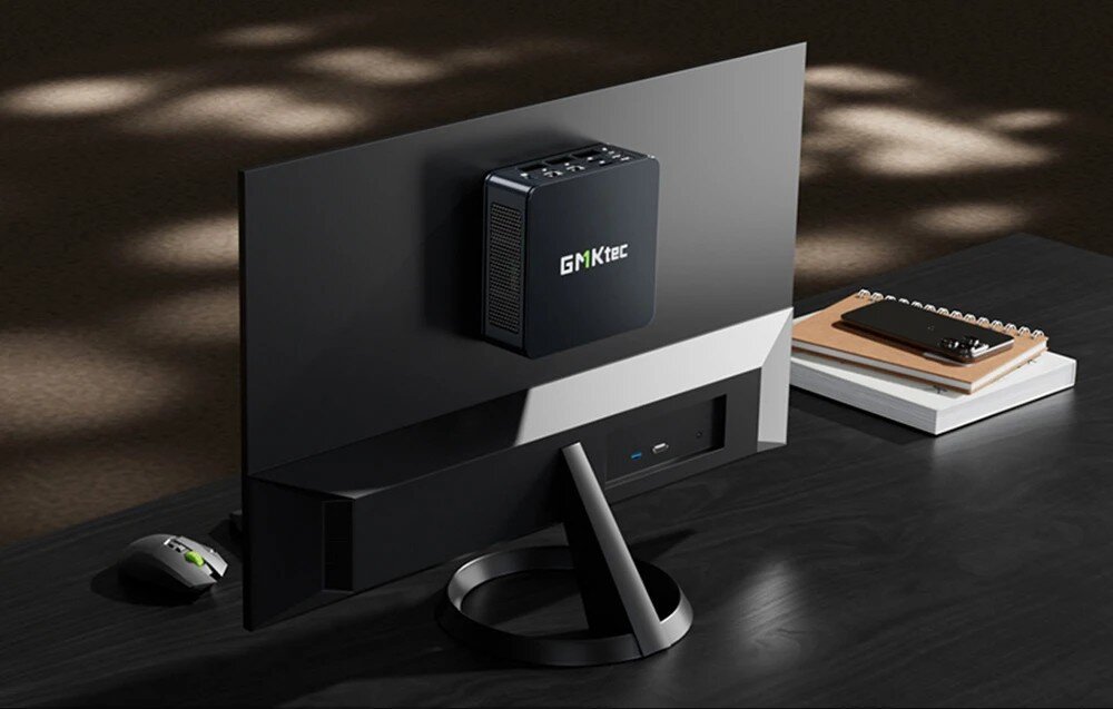 Komputer GMKTEC M6 Ultra Czarny mini komputer GMKtec ustawiony na biurku przy monitorze, z komputerową myszą po lewej stronie i notesami po prawej