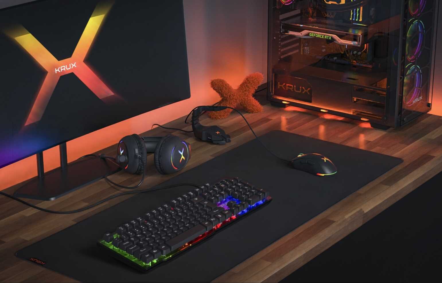 Na biurku znajduje się podkładka, klawiatura z podświetleniem RGB, mysz oraz słuchawki z logo KRUX. W tle widać monitor z logo KRUX oraz komputer z podświetleniem RGB i napisem KRUX na obudowie.